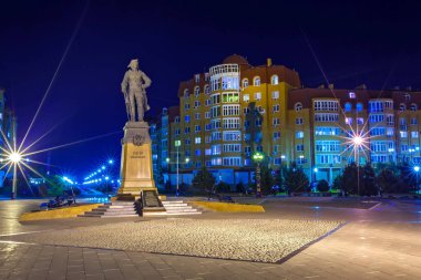 Monument to Great Peter in the darkness of the embankment on backdrop of urban modern residential buildings and walking alley illuminated by light of street lampposts. Night Astrakhan City, Russia. clipart