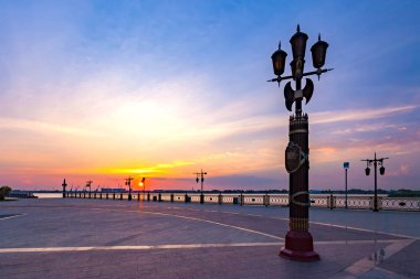 Volga nehir kıyısında günbatımı güzel bir renkli gökyüzü ve giden bir turuncu güneş sırasında önünde kare dekoratif sütun. Petrovskaya dolgu, Astrakhan, Rusya Federasyonu.