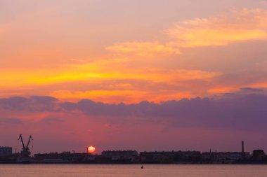 Büyük bir kırmızı güneş disk turuncu bulutların ötesinde şehir manzarası siluetleri evleri ve yükleme denizin içine ayarlar. Kıyı nehir günbatımı, Astrakhan, Rusya Federasyonu.