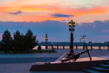 Çapa Volga nehir kıyısında günbatımı güzel bir renkli gökyüzü ve bir giden sırasında önünde kare üzerinde turuncu güneş ile dekoratif lamba direği. Petrovskaya dolgu, Astrakhan, Rusya Federasyonu.