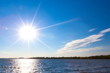 Bir büyük beyaz güneş disk ötesinde şehir manzarası ile küçük bulutların nehir dalgaları üzerinde güzel masmavi gökyüzünde ışık ışınları yayar. Kıyı Volga Nehri, Astrakhan, Rusya Federasyonu.