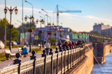 Fishing rods supported on a fence with stretched fishing line on the banks of the Volga River in the city with buildings and cranes on a blurred background. Petrovskaya embankment, Astrakhan, Russia. clipart