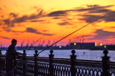 Uzun olta Volga Nehri üzerinde parlak renkli turuncu günbatımı üzerinde yalnız bir balıkçı yanında çite silüeti. Petrovskaya dolgu, Astrakhan, Rusya Federasyonu - Eylül 2014.