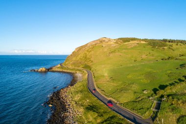 Northern Ireland, UK. Causeway Coastal Route a.k.a Antrim Coast Road near Ballygalley Head and resort with red cars. One of the most scenic coastal roads in Europe. Aerial view. clipart