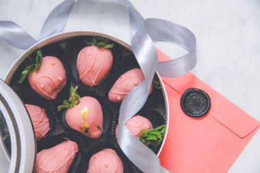 Şeffaf kapaklı bir hediye kutusunda çikolata kaplı çilekler, gri bir kurdele ve mühürlü balmumuyla kaplı bir zarf.