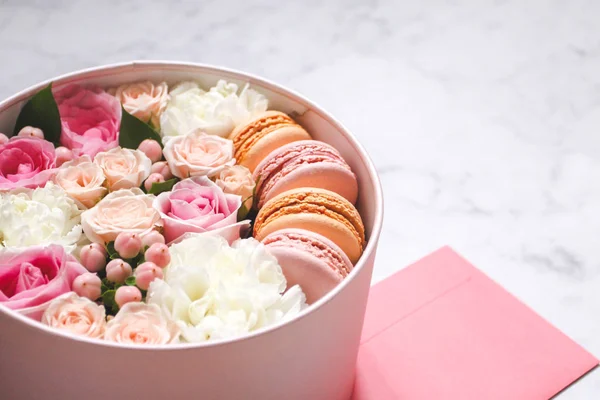 Gift round box with flowers, roses and macaroons almond cake with pink envelope on the table