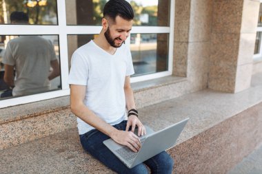 Güzel şık adam portresi bir korkuluk, cam bina karşı sokakta bir dizüstü bilgisayar ile çalışır üzerinde oturan beyaz tişört giymiş,
