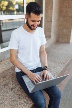 Güzel şık adam portresi bir korkuluk, cam bina karşı sokakta bir dizüstü bilgisayar ile çalışır üzerinde oturan beyaz tişört giymiş,