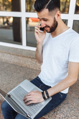 Bir parapetin üzerinde oturan beyaz bir t-shirt giyen bir güzel şık adam portresi ile bir dizüstü bilgisayar çalışır ve bir cam bina önünde sokakta telefonda konuşuyor,