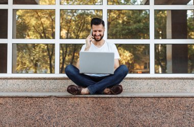 Bir parapetin üzerinde oturan beyaz bir t-shirt giyen bir güzel şık adam portresi ile bir dizüstü bilgisayar çalışır ve bir cam bina önünde sokakta telefonda konuşuyor,