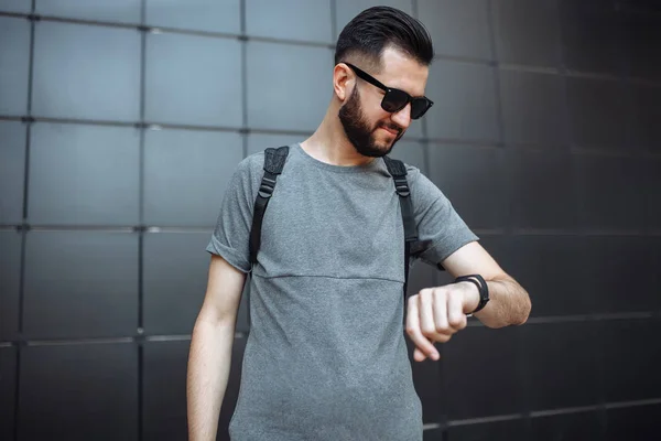 Güzel şık tekiyle saate benzeyen hipster gözlüklü, portre siyah duvar arka plan üzerinde duran bir gri boş t-shirt giymiş.