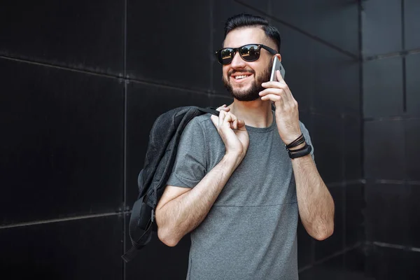 Portrait of a beautiful stylish guy, hipster with glasses, talking on the phone, dressed in a gray empty t-shirt, standing on a black wall background.