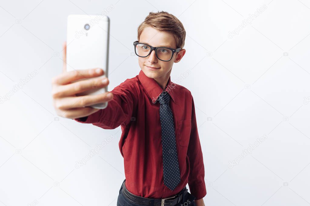 Portrait of a positive and emotional schoolboy, takes a selfie on your smartphone, white background, glasses, red shirt, business theme,