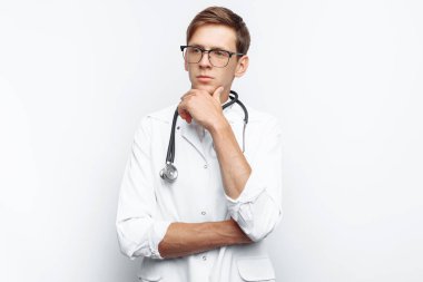 Düşünceli görünüm stajyer stetoskop bir boyun ile stüdyoda, gösterir beyaz bir arka plan üzerinde genç bir doktor portresi