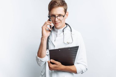 Genç ve yakışıklı doktor hasta ile telefon, stajyer öğrenci klasörle elinde, konuşurken beyaz arka plan,