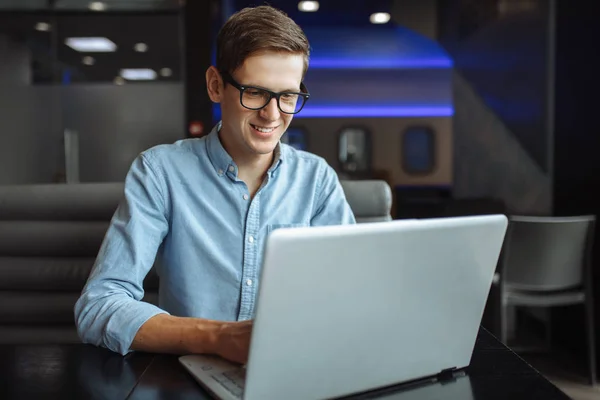 Portrait of a young man with a good mood, a businessman in a shirt and glasses, who works on a laptop in a cafe,