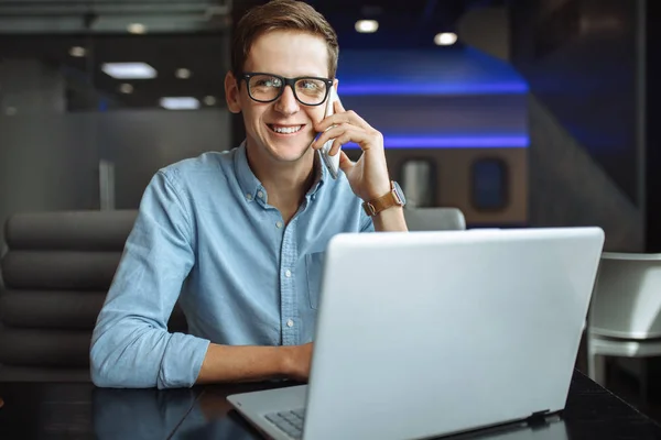 Portrait of a young man with a good mood, a businessman in a shirt and glasses, who works on a laptop and talking on the phone in a cafe,