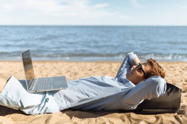 Gözlüklü, Genç adam tatilde çalışma karşı deniz, plaj onun laptop üzerinde kum üzerinde yalan,