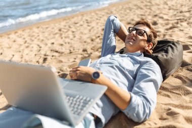 Gözlüklü, Genç adam tatilde çalışma karşı deniz, plaj onun laptop üzerinde kum üzerinde yalan,
