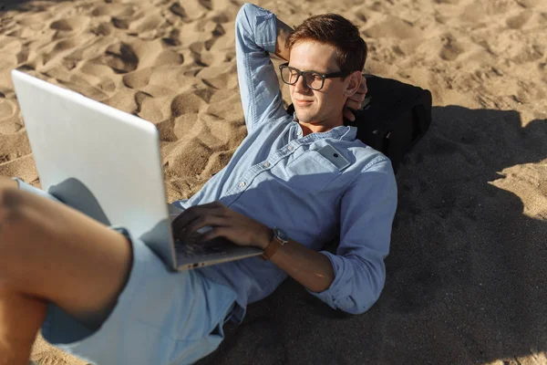 Gözlüklü, Genç adam tatilde çalışma karşı deniz, plaj onun laptop üzerinde kum üzerinde yalan,