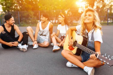 Bir grup arkadaşıyla birlikte bir skate Park, yakın çekim güzel gülümseyen kız bir grup arkadaşıyla birlikte, arka plan üzerinde asfalt üzerinde gün batımında eğleniyor gitar, iyi yaz ruh hali