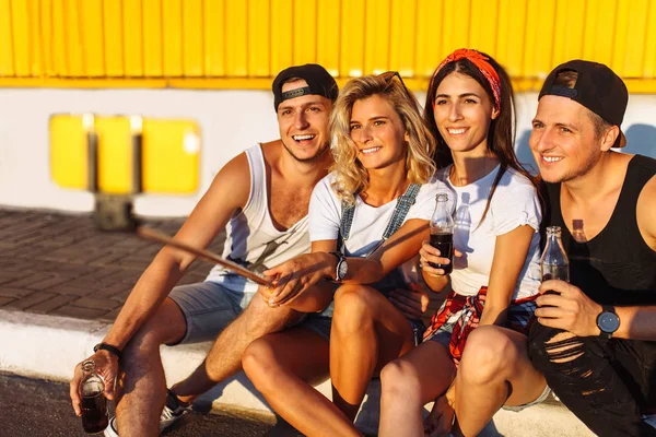 A group of friends makes a selfie sitting on the pavement, the company of young people taking pictures with a selfie stick, cheerful company, Sunny day