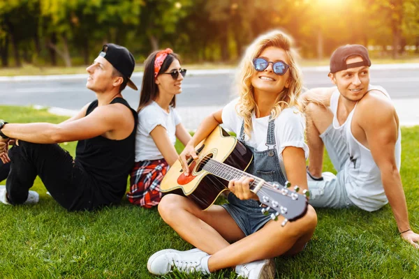 Company of friends having fun at sunset sitting on the grass, close-up beautiful smiling girl on the background of a group of friends, playing guitar, good summer mood