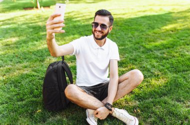 şık hipster öğrenci Park'ın bahçesinde oturan beyaz gömlekli ve smartphone bir selfie alır