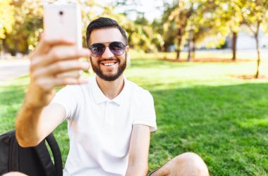 şık hipster öğrenci Park'ın bahçesinde oturan beyaz gömlekli ve smartphone bir selfie alır