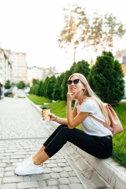 Kahve ile açık havada beyaz bir t-shirt giyen güneş gözlüğü, giyen genç bir kadın portresi. Çimenlerin üzerine oturmuş ve kahve içme kız.