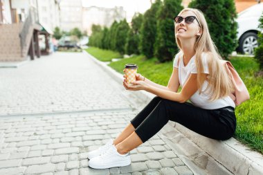 Kahve ile açık havada beyaz bir t-shirt giyen güneş gözlüğü, giyen genç bir kadın portresi. Çimenlerin üzerine oturmuş ve kahve içme kız.