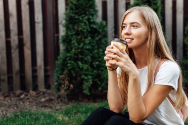 Genç bir adam portresi bir fincan kahve ile açık havada beyaz tişört giymiş. Çimenlerin üzerine oturmuş ve kahve içme kız.