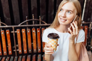 Evet ve beyaz tişört, ahşap bir yaprak döken duvara bir bankta oturmuş, bir fincan kahve içme genç güzel kız