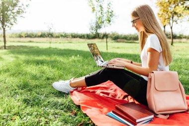 Bir dizüstü bilgisayarda çalışan bir güzel kız öğrencinin gözlüklü, görüntü üzerinde ödev Park kırmızı bir battaniyeye oturmuş