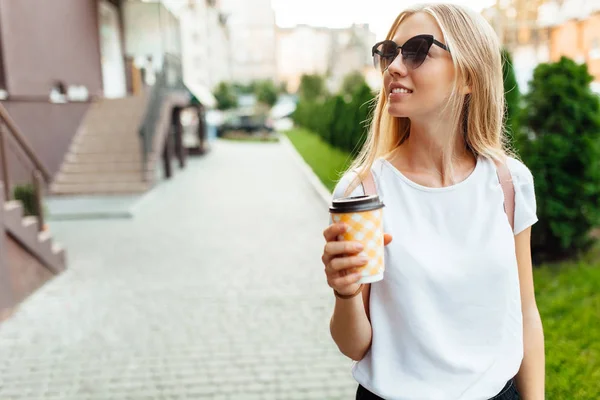 Açık havada beyaz bir t-shirt giyen ve bir fincan kahve tutan güneş gözlüğü, giyen genç bir kadın portresi. Şehirde yürüyüş ve kahve içme kız.