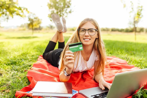 美丽的女孩在眼镜与膝上型电脑吹捧一张信用卡坐在公园的毯子上 — 图库照片
