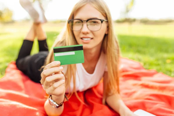 美丽的女孩与眼镜显示信用卡坐在公园的毯子 — 图库照片
