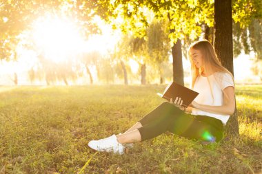 güzel kız ağaca bir Parkı günbatımında oturan bir kitap okuma