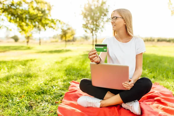 Beautiful girl in glasses with laptop touts a credit card sitting in the Park on the blanket