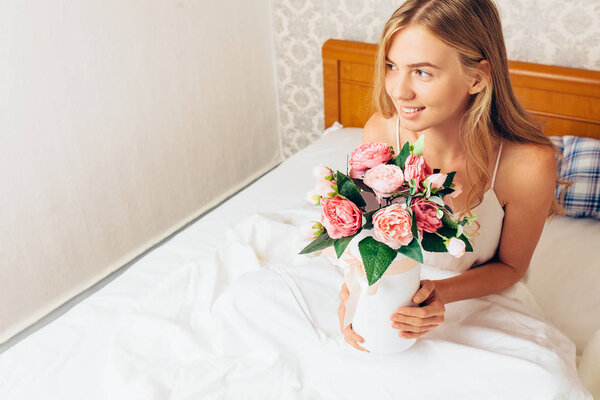 Young beautiful girl with peony flowers, sitting on the bed. She just woke up and was given a bouquet of flowers