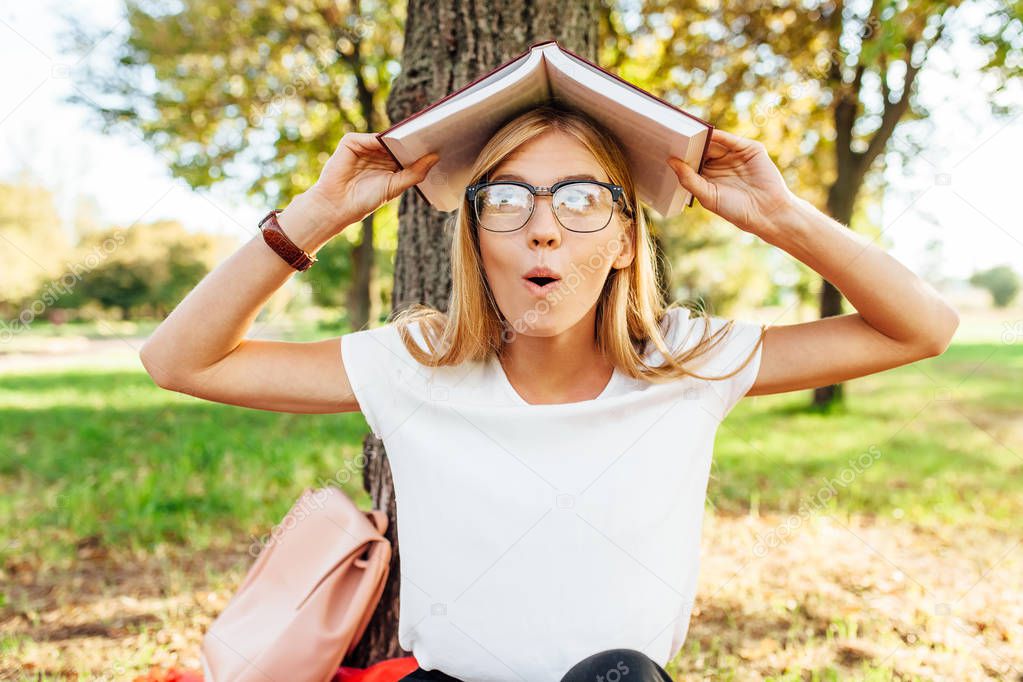 Young positive student with glasses, funny posing with a book on his head, in the Park sitting on a red blanket