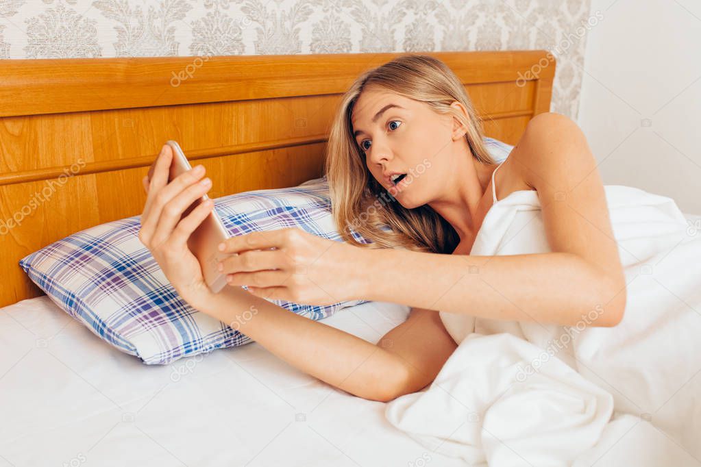 A young sleepy girl in shock, with her eyes wide open in her mouth, lying in bed, looks at the smartphone and realizes that she is late for the robot