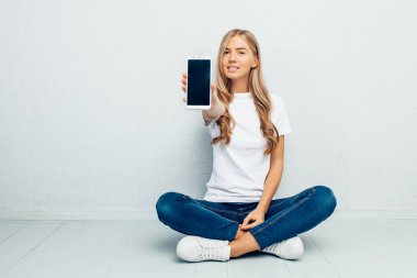 Beyaz bir t-shirt giyen güzel bir genç kız bacaklarını geçti bir boş cep telefonu ekran gri bir arka plan üzerinde gösterilen yere oturuyor
