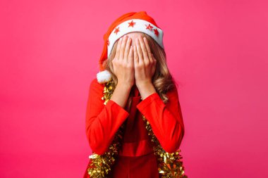 Kız Noel Baba'nın kırmızı şapkalı ve boynunda, gelin teli ile kırmızı bir arka plan üzerinde birinden saklanıyor ellerini, gözlerini kapatır