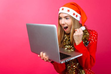 Mutlu kız kırmızı kazak ve bir laptop arıyorum Santa şapka heyecanlı hakkında good news, şok kız gösterir jest kırmızı bir arka plan üzerinde başarı zafer. Noel iş kavramı