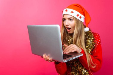 Şok kız kırmızı kazak ve Noel Baba şapkası, elinde, bir dizüstü bilgisayar ile dizüstü bilgisayar ekranında, kırmızı bir arka plan üzerinde sürpriz bakar