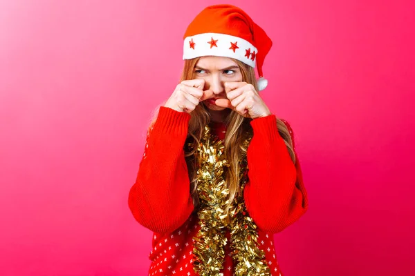 Muchacha molesta en sombrero de Santas y con oropel en el cuello, se frota los ojos y quiere llorar con resentimiento sobre un fondo rojo . — Foto de Stock