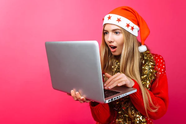 Shocked girl in a red sweater and Santa Claus hat, with a laptop in hand, looks in surprise at the laptop screen, on a red background — Stock Photo, Image