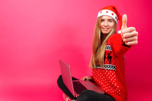 Gelukkig meisje in de rode trui en kerstmuts, zitten met laptop en shows klasse op de camera, op rode achtergrond. Kerstmis, vakantie, bedrijfsconcept — Stockfoto