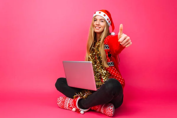 Šťastná dívka v Santa čepice, sedí s třídou laptop a ukazuje na kameru, na červeném pozadí a červený svetr. — Stock fotografie